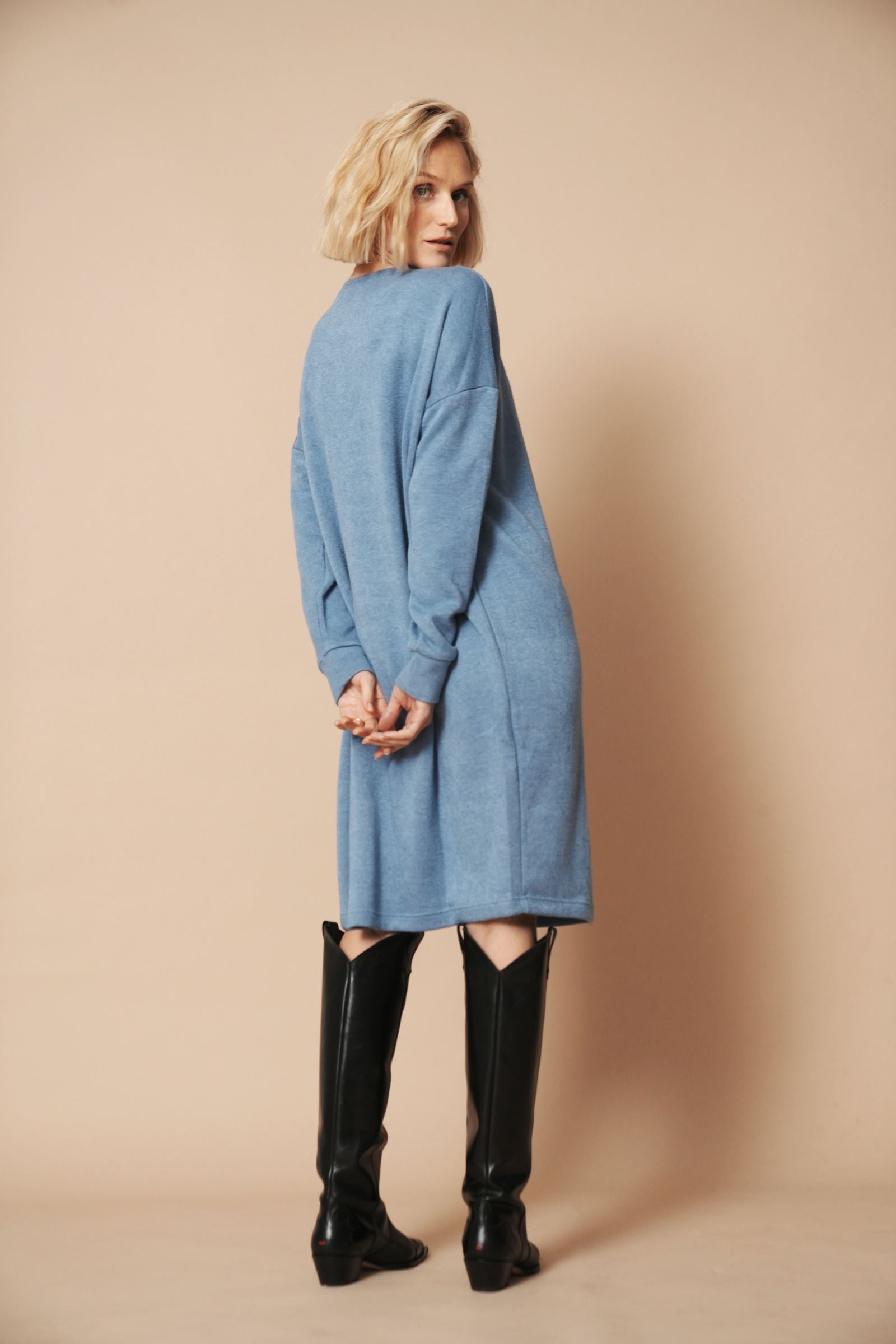 Das hellblaue Strickkleid mit langen Ärmeln wird von einer Frau mit blonden Haaren getragen. Das Outfit wird von hinten gezeigt.