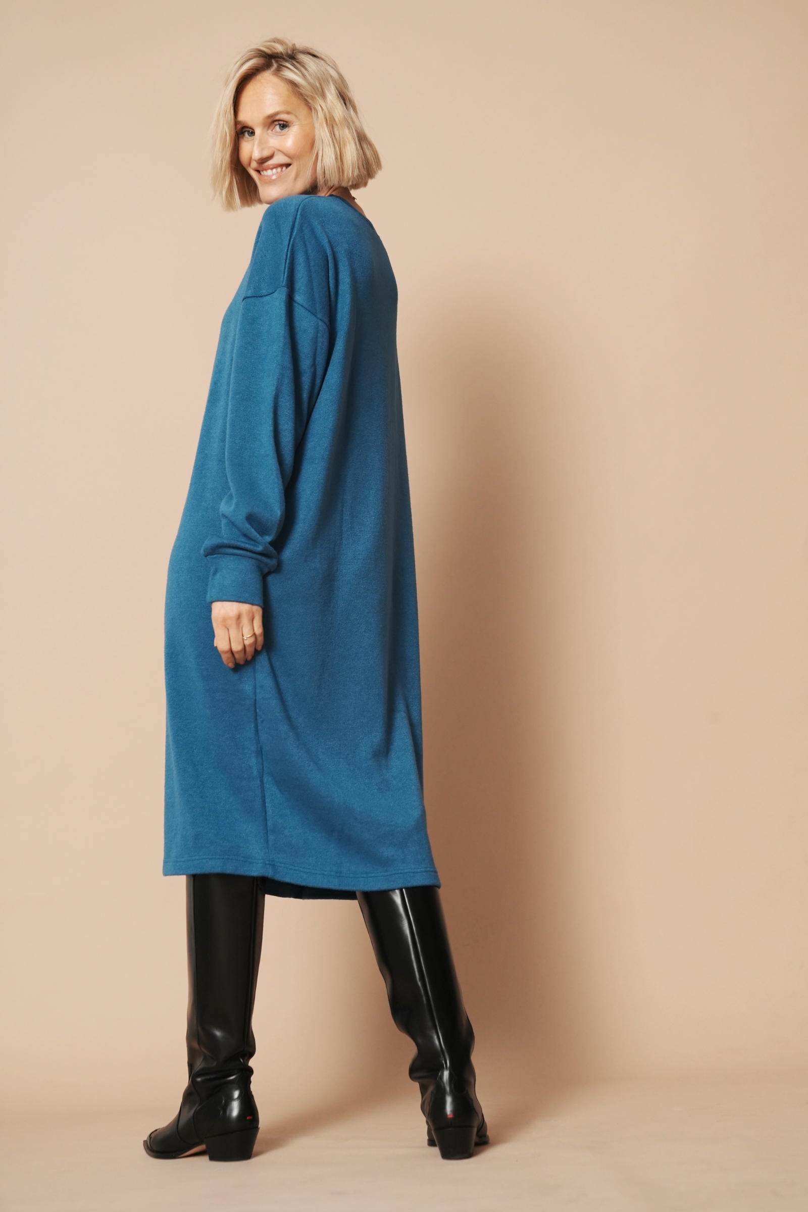 Das Strickkleid in petrol mit langen Ärmeln wird von einer Frau mit blonden Haaren getragen. Das Outfit wird von der linken Seite gezeigt.