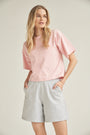 Ein blondes Model trägt eine Baumwolle-Shorts in der Farbe grau. Sie kombiniert das Outfit mit einem Shirt in rosa. Das Model steht vor einem hellen Hintergrund und ihre Hände befinden sich in den Seitentaschen der kurzen Hose.