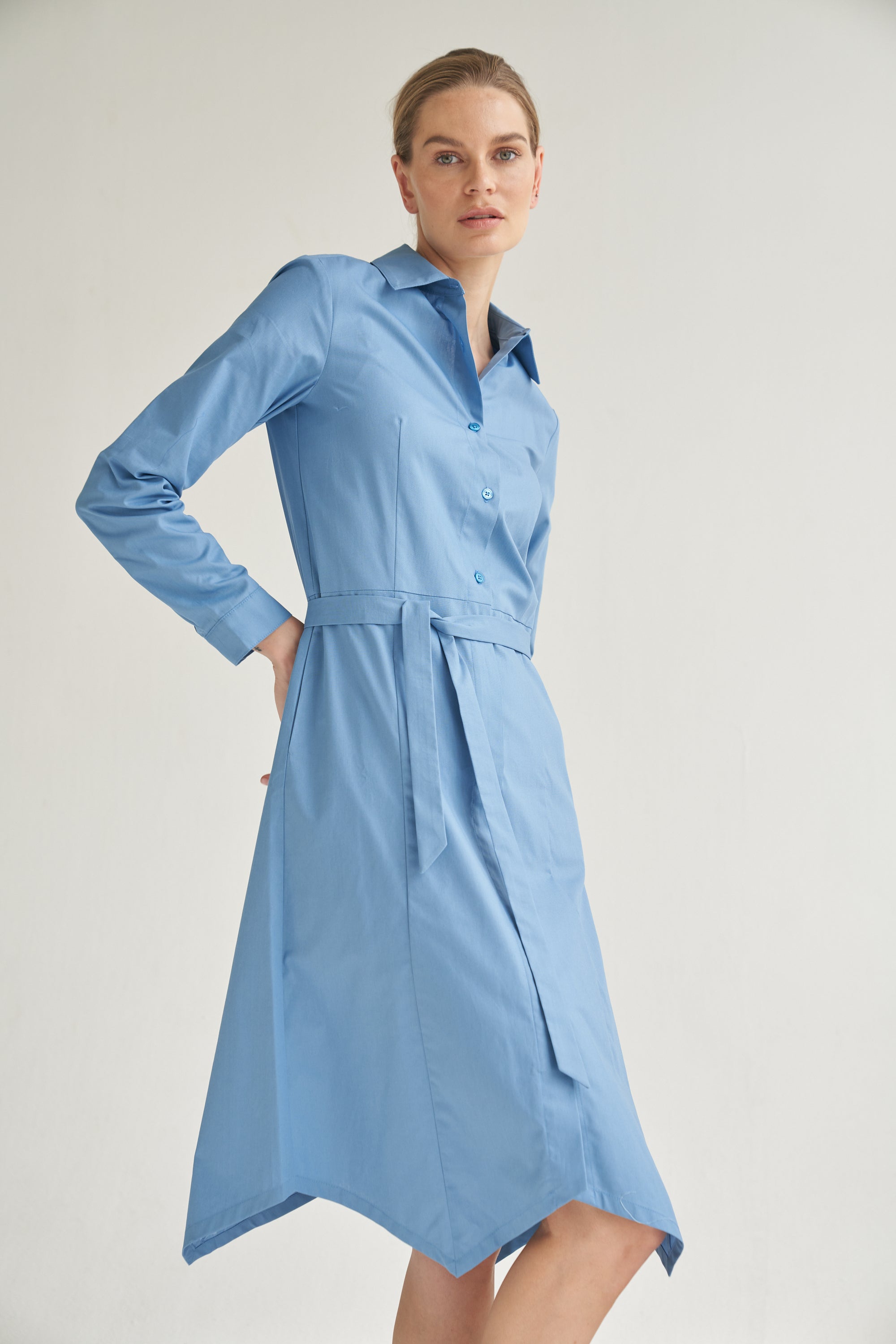 Unser Model trägt ein Hemdblusenkleid in blau von Bellasor. Das Model hat blondes Haar und Sie zeigt das elegante Outfit seitlich von vorne.