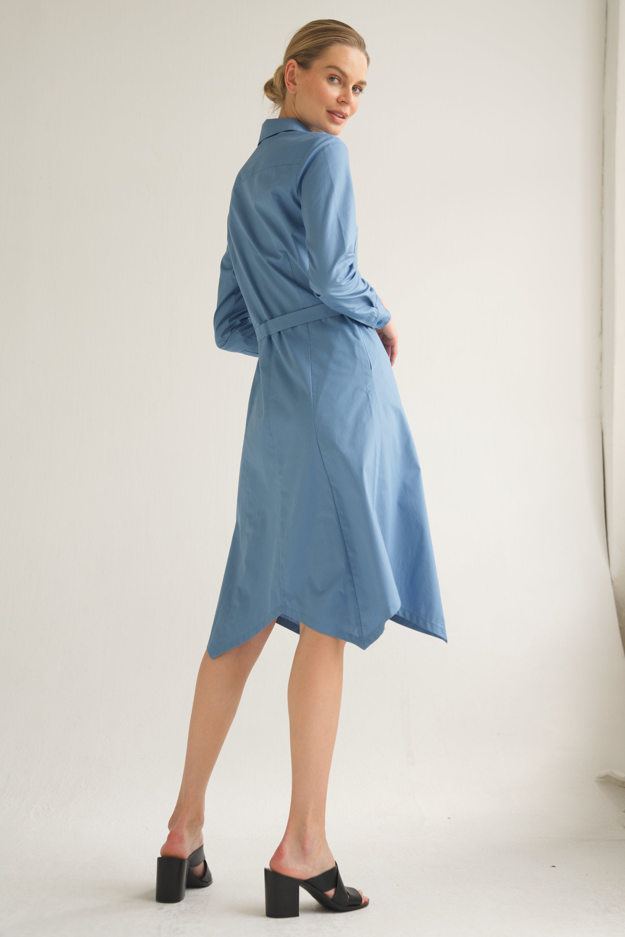 Unser Model trägt ein Hemdblusenkleid in blau von Bellasor. Das Model hat blondes Haar und Sie zeigt das elegante Outfit seitlich von hinten.