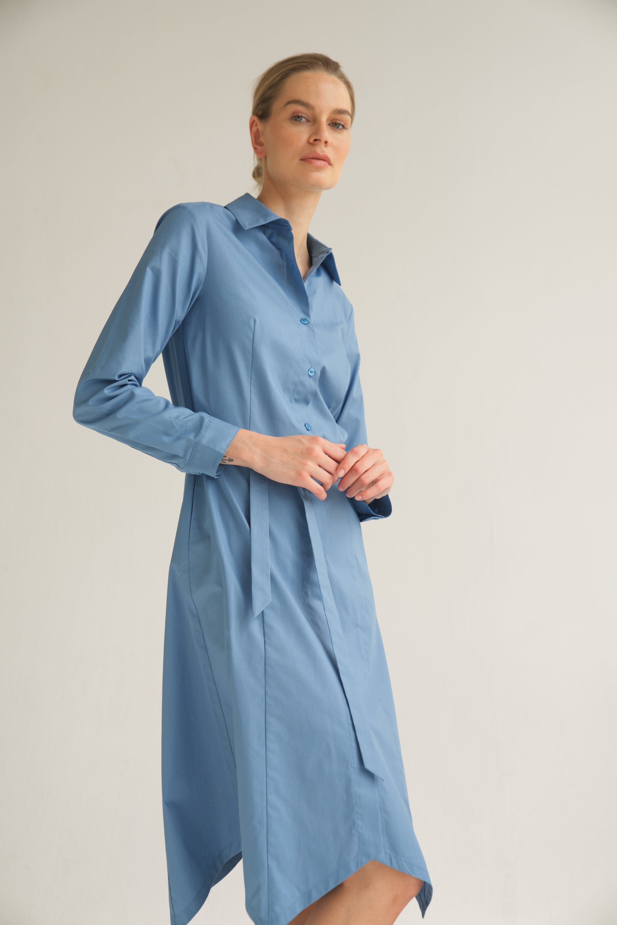 Unser Model trägt ein Hemdblusenkleid in blau von Bellasor. Das Model hat blondes Haar und Sie zeigt das elegante Outfit seitlich von vorne.