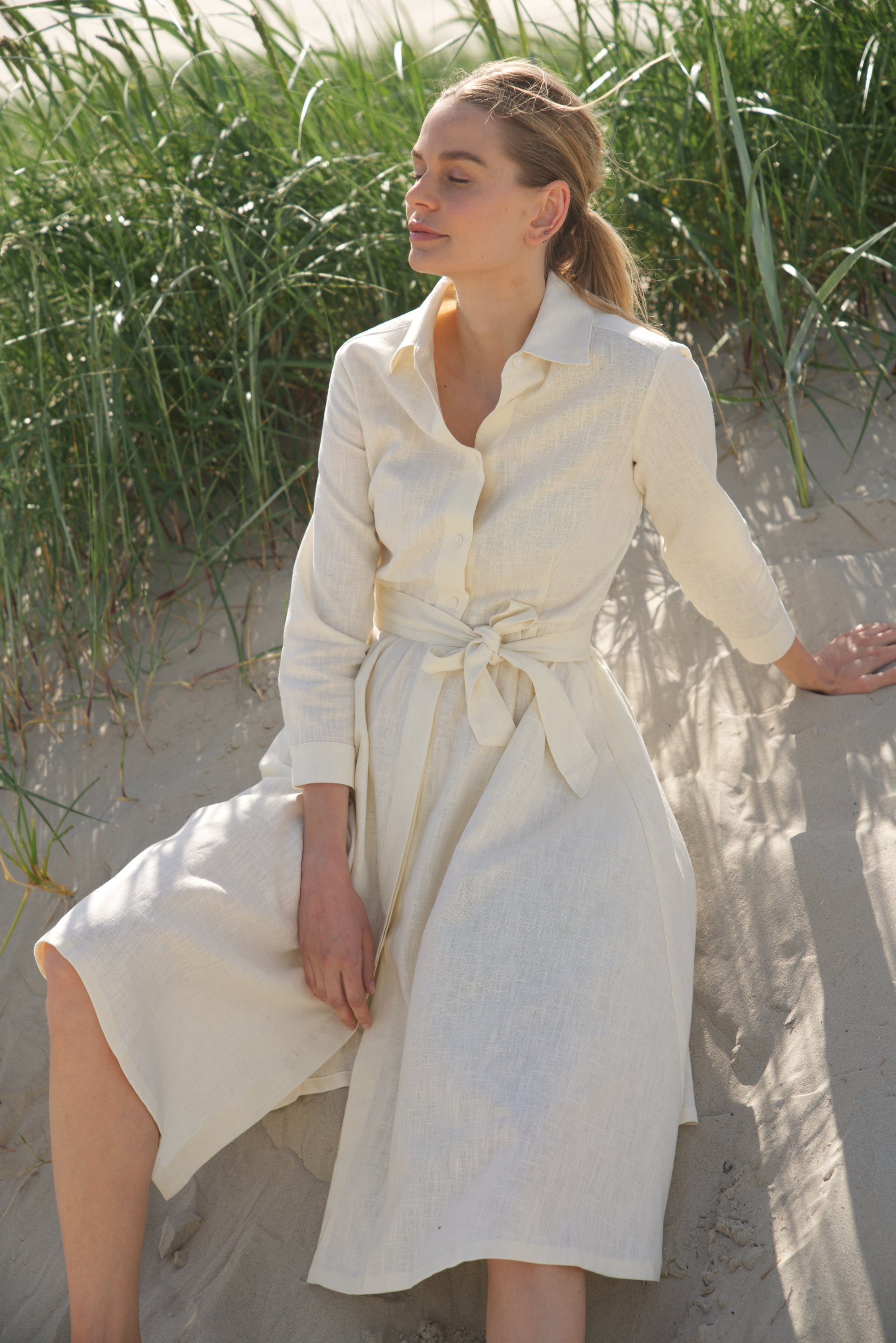 Unser Leinenkleid in der Farbe cremeweiss von der Marke Bellasor wird von einem blonden Model getragen. Sie sitzt bei Sonnenschein in den Dünen von Sankt Peter Ording.