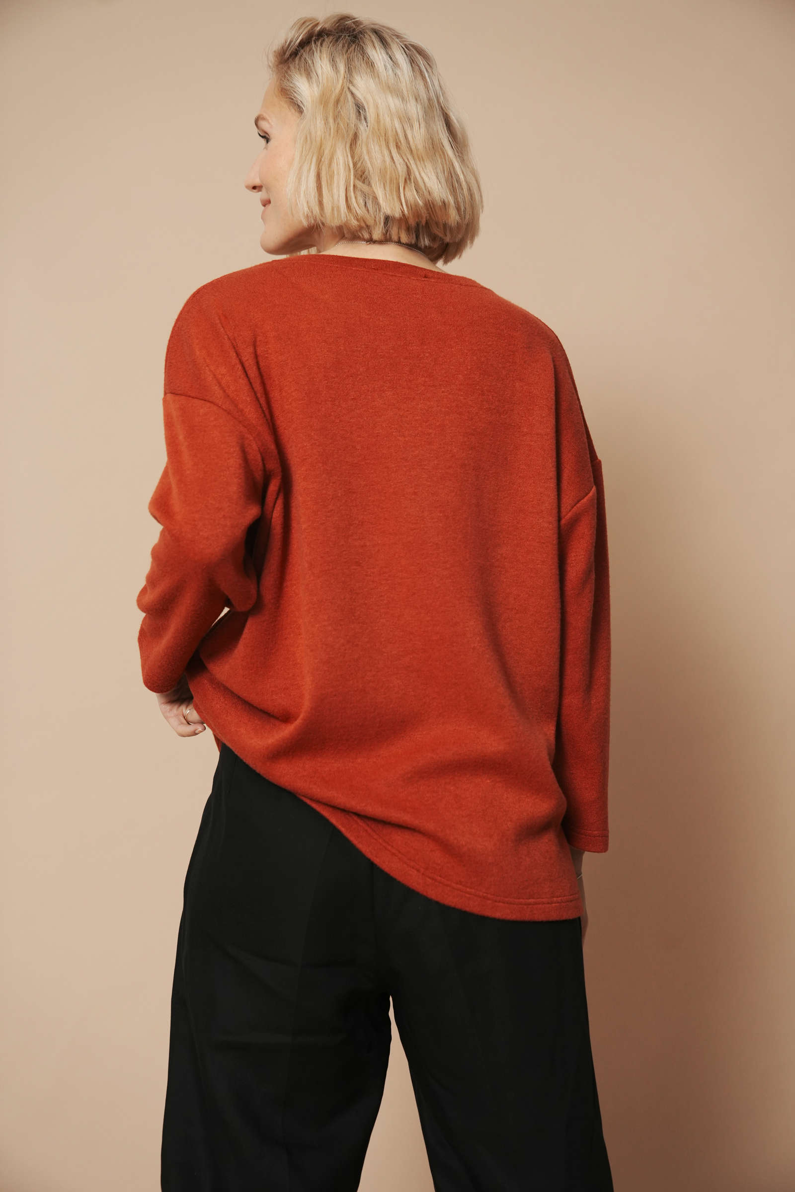 Blondes Model trägt einen hochwertigen Strickpullover für Damen. Sie zeigt die Ansicht von hinten und hat dabei die linke hand am Pullover und rafft ihn etwas nach oben.