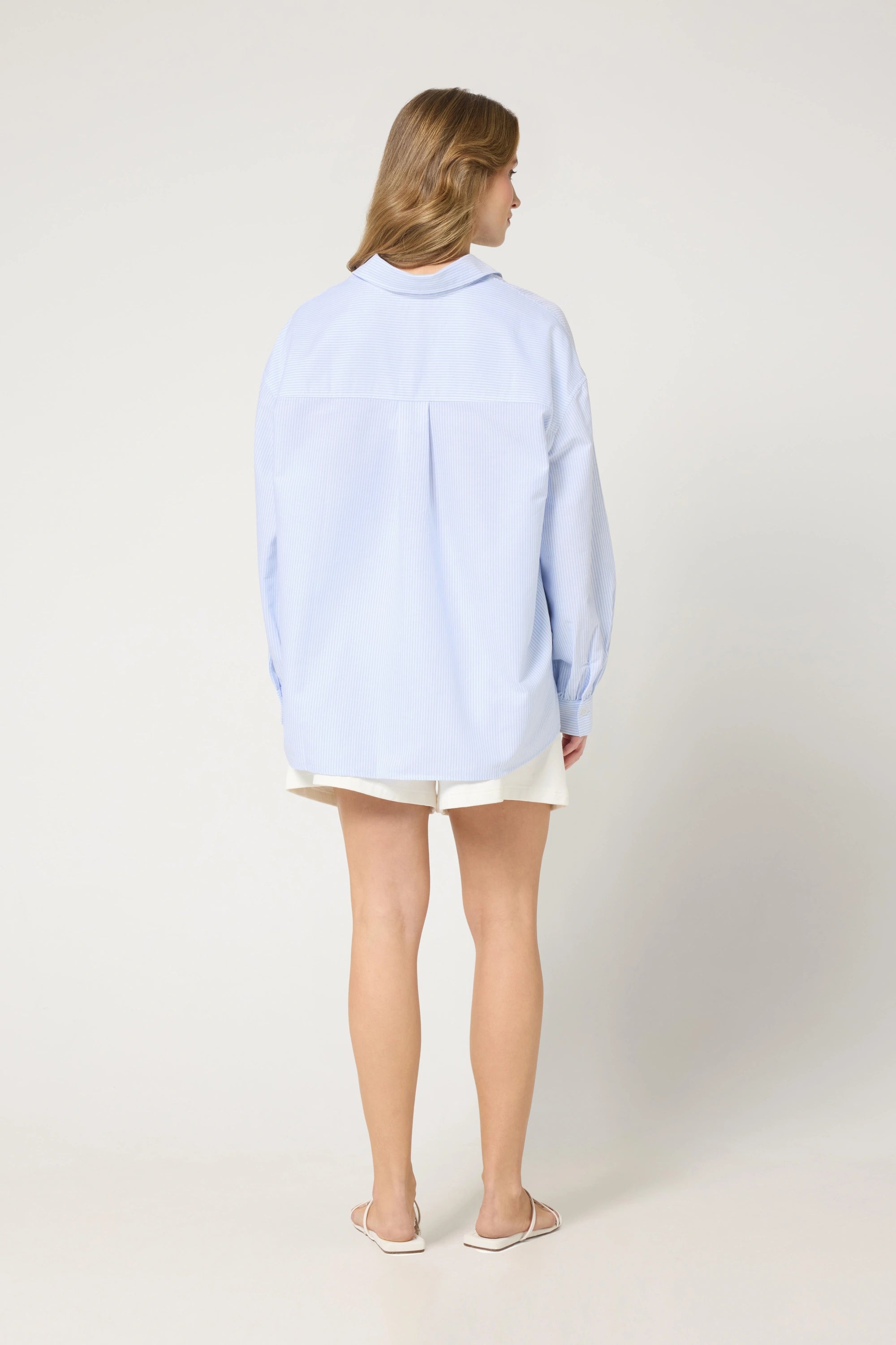 Rückansicht der hellblauen, gestreiften Oversize-Bluse für Damen, getragen von einem weiblichen Model im Studio vor einer cappuccino-farbenen Wand.