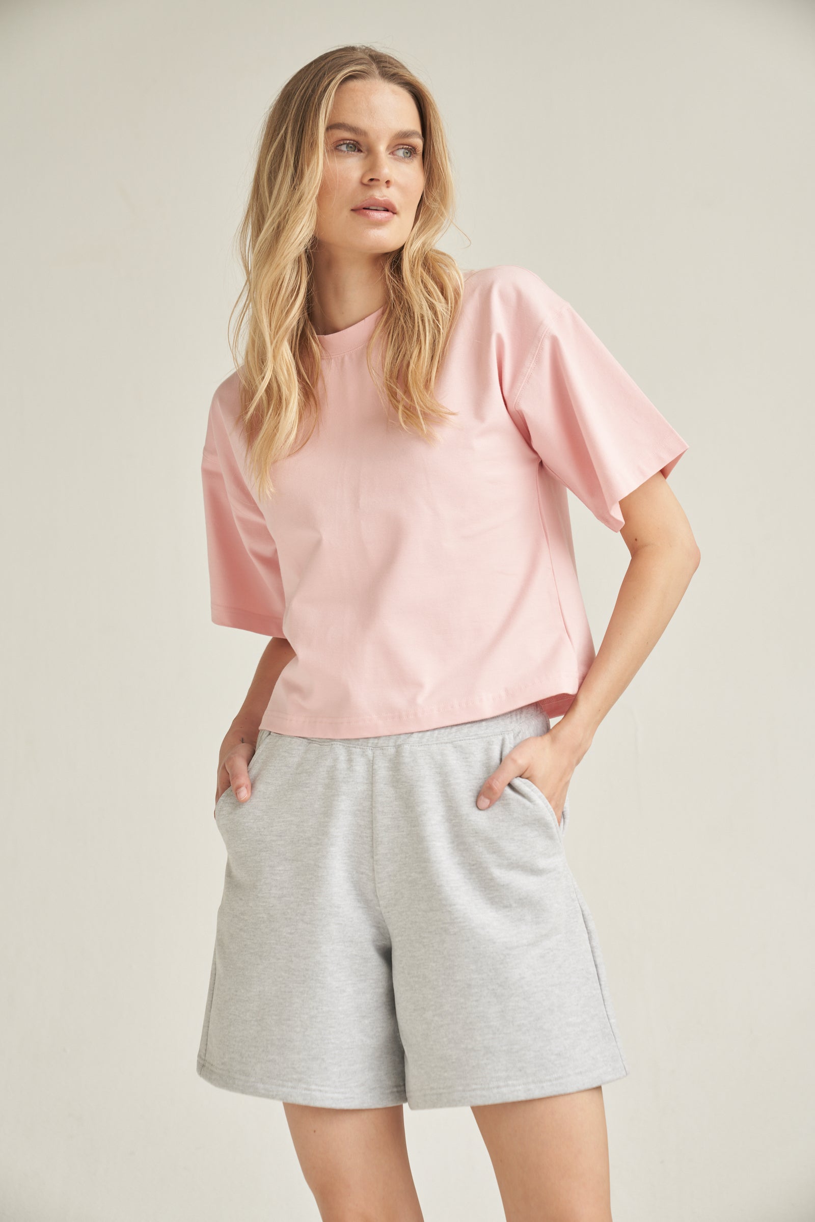Ein blondes Model trägt eine Baumwolle-Shorts in der Farbe grau. Sie kombiniert das Outfit mit einem Shirt in rosa. Das Model steht vor einem hellen Hintergrund und ihre Hände befinden sich in den Seitentaschen der kurzen Hose.