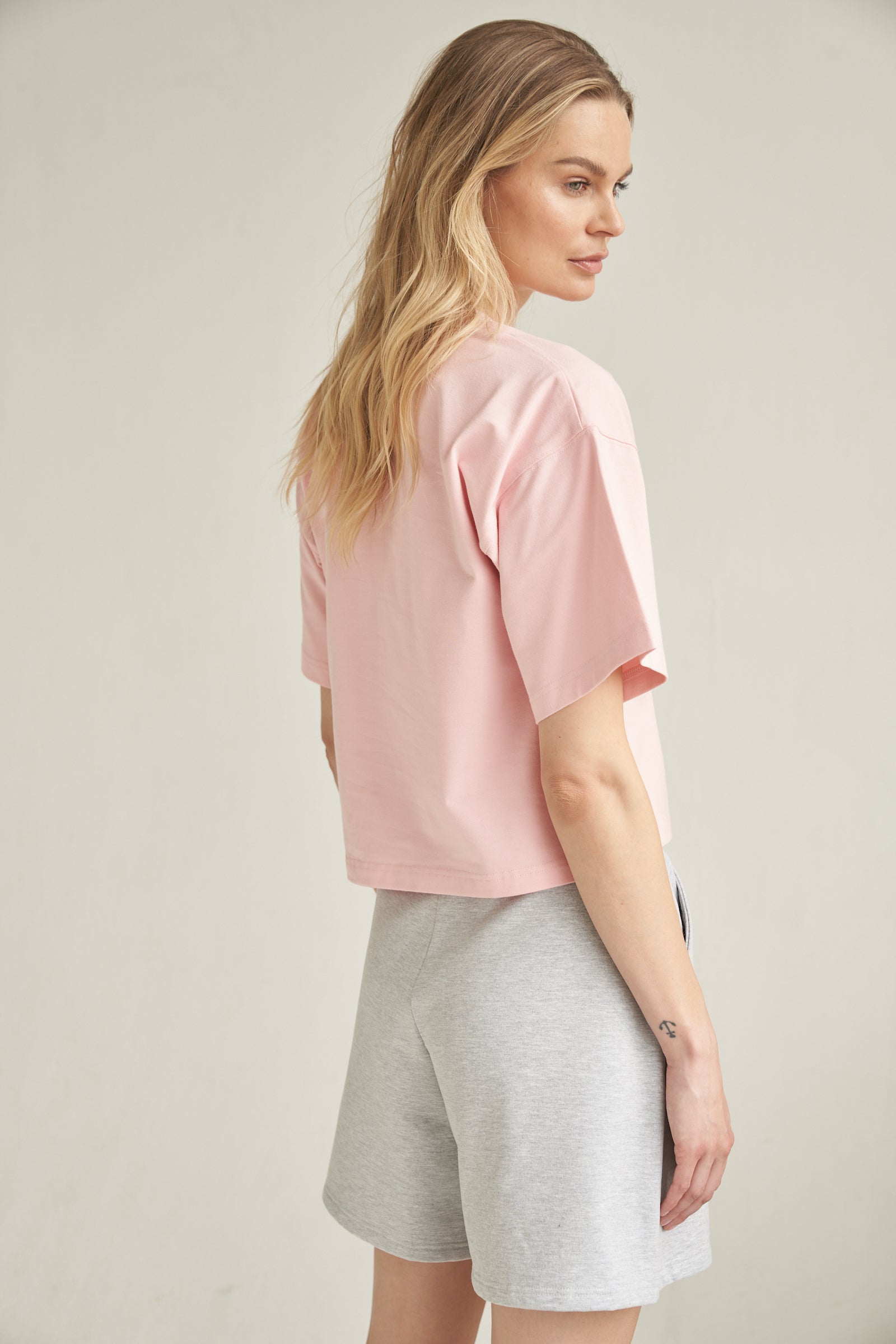 Ein blondes Model trägt eine Baumwoll-Shorts in der Farbe grau. Sie kombiniert das Outfit mit einem Shirt in rosa. Das Model steht seitlich vor einem hellen Hintergrund. Der Rücken ist zu erkennen und ihre Hände befinden sich in den Seitentaschen der kurzen Hose.