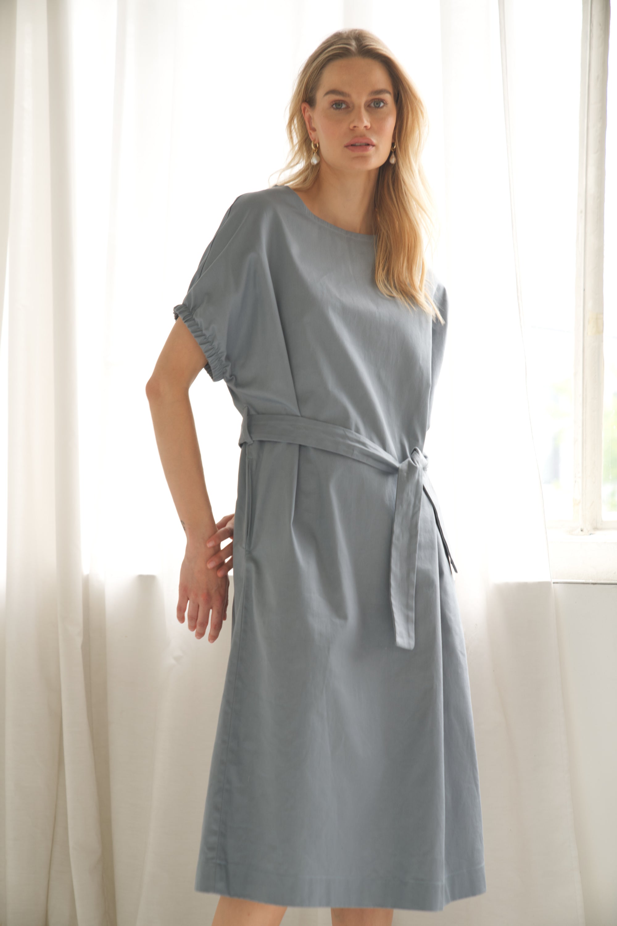 Oversize-Kleid aus Baumwolle in der Farbe blau wird von einem blonden Model getragen. Sie zeigt die Ansicht von vorne und die Arme sind hinter dem Rücken verschränkt.