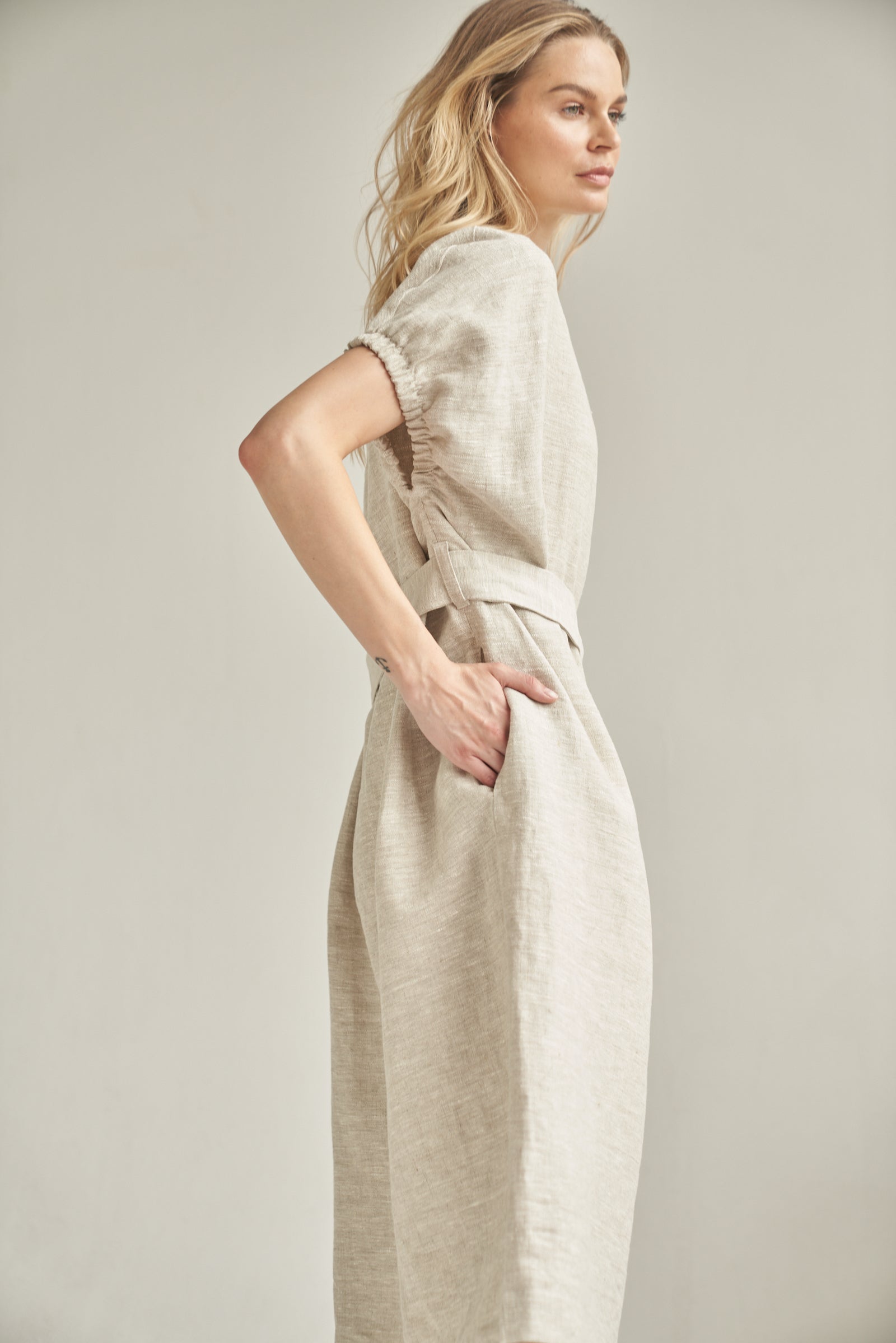 Das Oversize-Kleid aus Leinen in der Farbe natur wird von einem blonden Model getragen. Sie steht seitlich vor einem hellen Hintergrund und hat ihre rechte Hand in der integrierten Eingrifftasche.