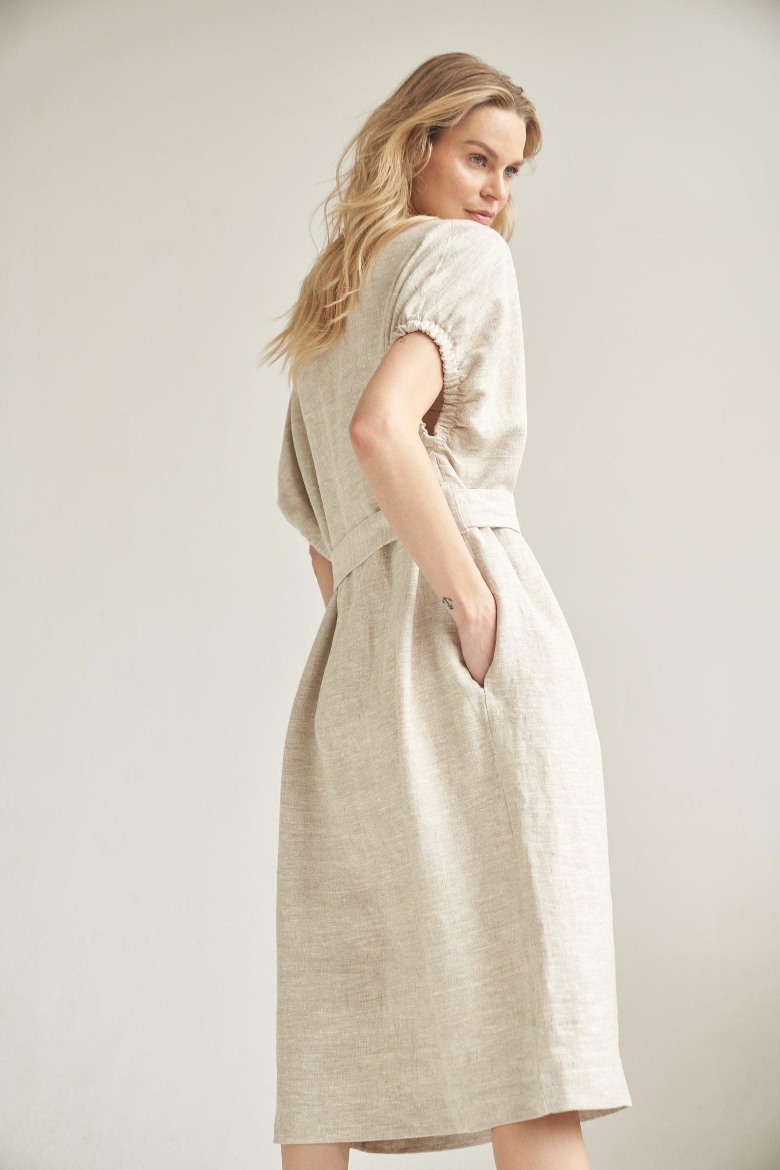 Das Oversize-Kleid aus Leinen in der Farbe natur wird von einem blonden Model getragen. Sie steht vor einem hellen Hintergrund und hat ihre rechte Hand in der integrierten Eingrifftasche. Die Rückseite des Oversize-Kleid ist leicht von der Rückseite zu sehen.
