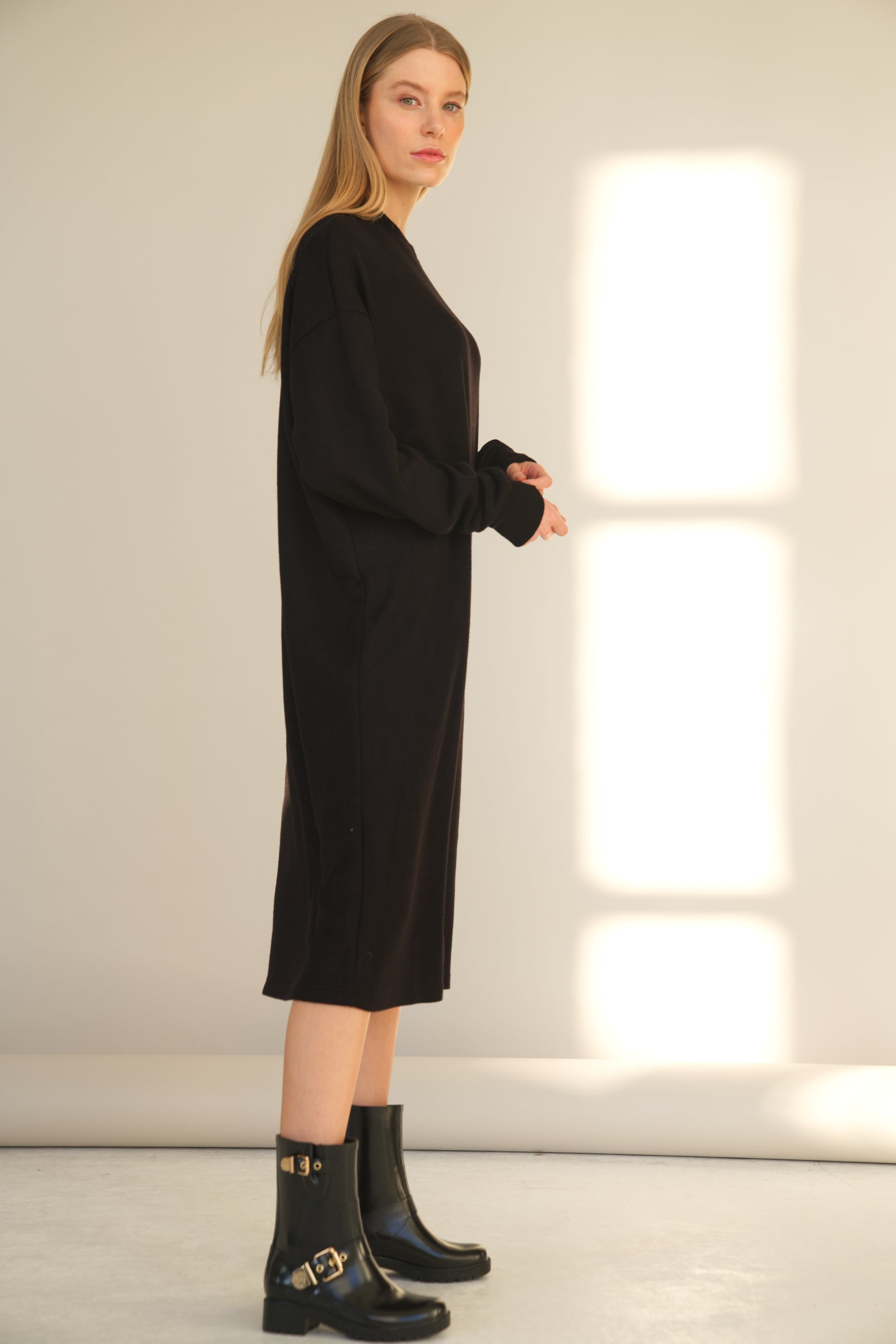 Unser Model trägt ein Strickkleid mit langarm in schwarz von Bellasor. Das Model ist von der Seite zu sehen und trägt schwarze Stiefel.