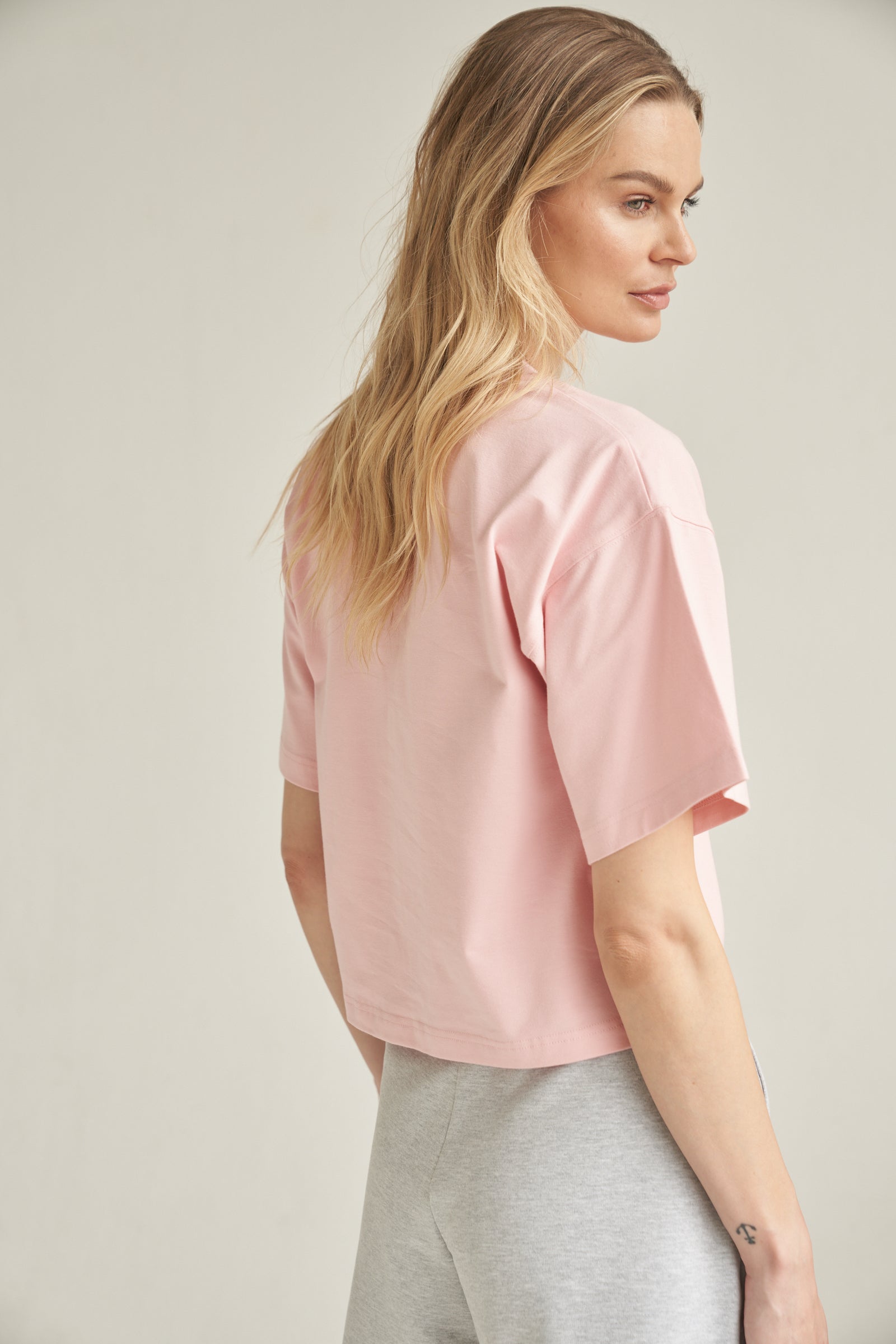 Unser T-Shirt aus Bio-Baumwolle in der Farbe rosa von der Marke Bellasor wird von einem blonden Model getragen. Sie steht vor einem hellen Hintergrund und zeigt das T-Shirt seitlich von hinten. Sie trägt dazu eine hellgraue Short von Bellasor.