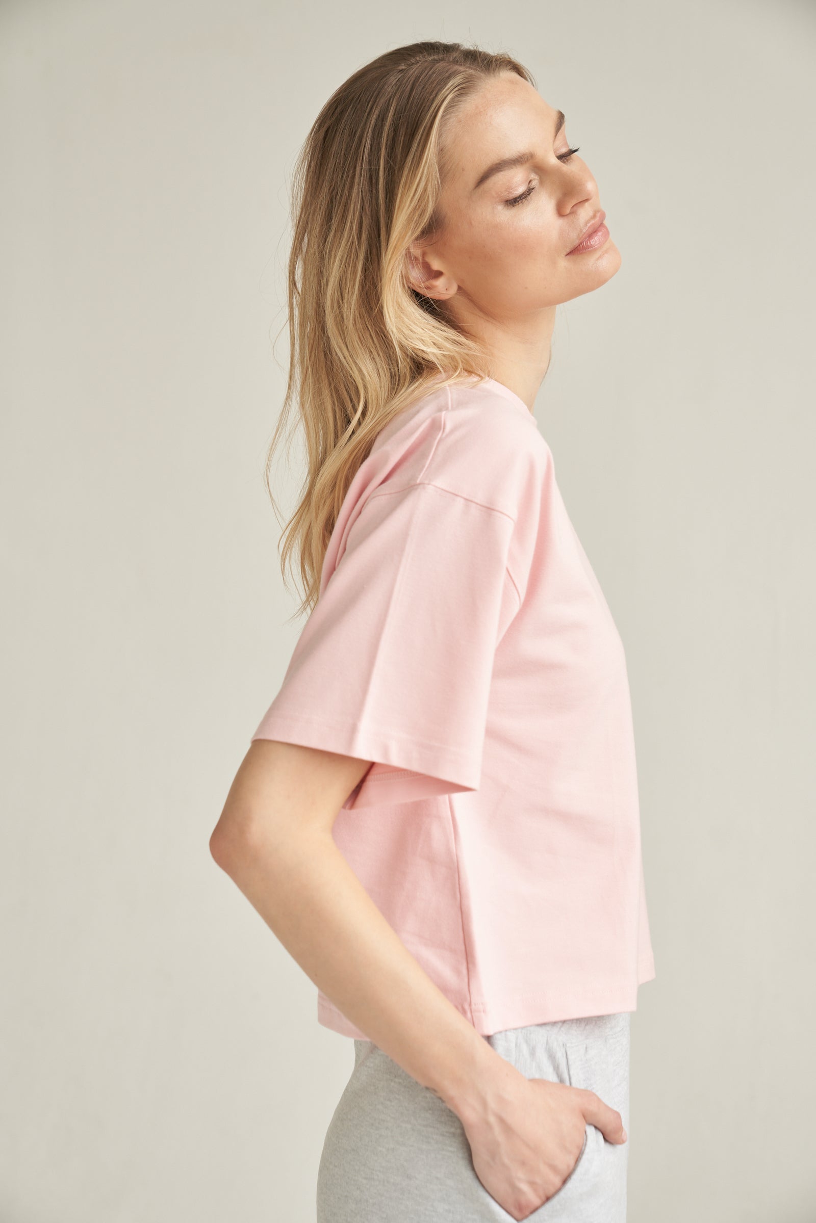 Unser T-Shirt aus Bio-Baumwolle in der Farbe rosa von der Marke Bellasor wird von einem blonden Model getragen. Sie steht vor einem hellen Hintergrund und zeigt das T-Shirt von der rechten Seite. Ihre rechte Hand steckt in der Tasche einer hellgrauen Short von Bellasor.