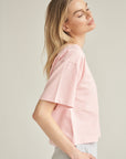Unser T-Shirt aus Bio-Baumwolle in der Farbe rosa von der Marke Bellasor wird von einem blonden Model getragen. Sie steht vor einem hellen Hintergrund und zeigt das T-Shirt von der rechten Seite. Ihre rechte Hand steckt in der Tasche einer hellgrauen Short von Bellasor.