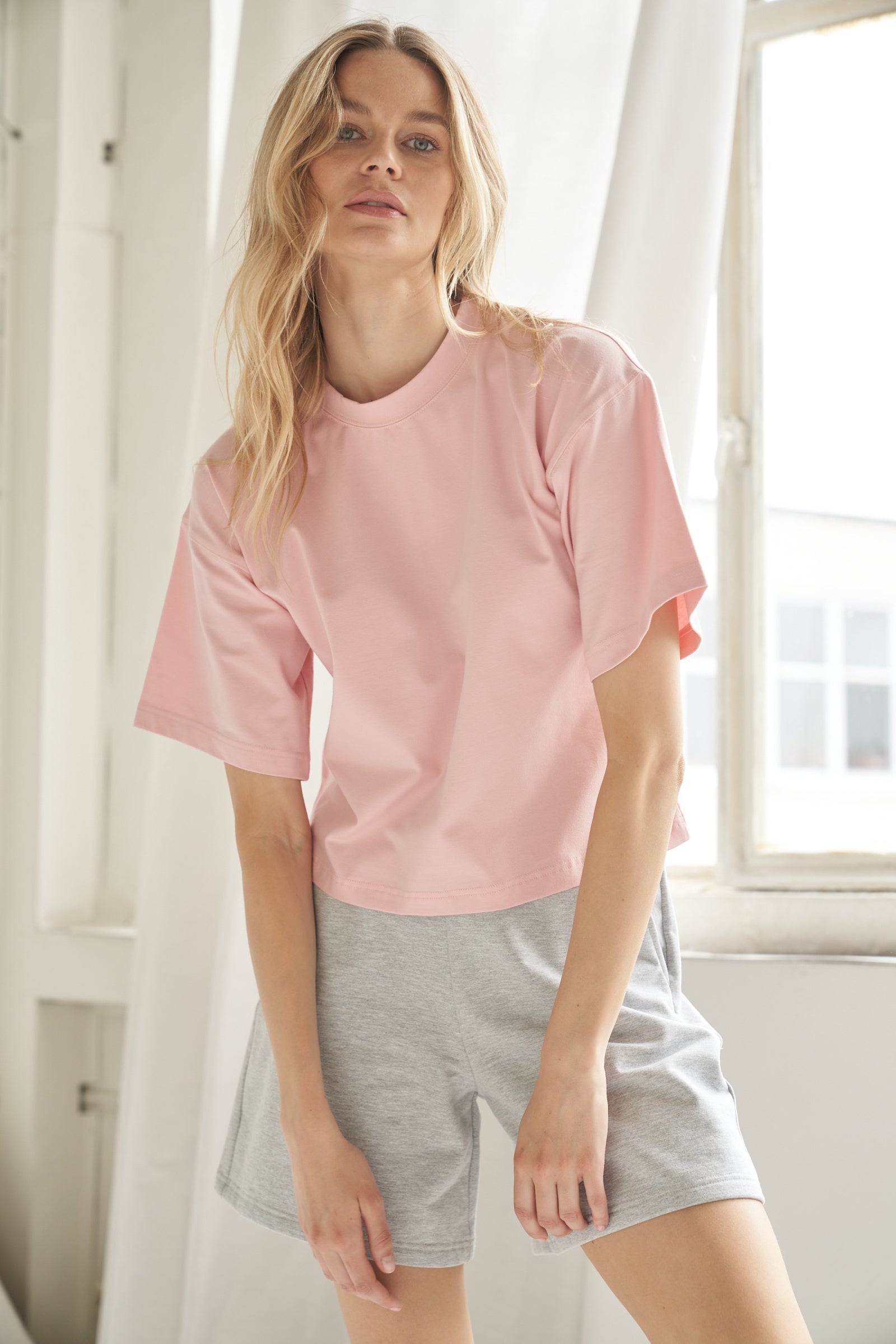 Unser T-Shirt aus Bio-Baumwolle in der Farbe rosa von der Marke Bellasor wird von einem blonden Model getragen. Sie zeigt das T-Shirt von vorne, befindet sich in einem hellen Raum und das Sonnenlicht scheint durch die Fenster im Hintergrund.