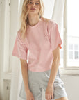 Unser T-Shirt aus Bio-Baumwolle in der Farbe rosa von der Marke Bellasor wird von einem blonden Model getragen. Sie zeigt das T-Shirt von vorne, befindet sich in einem hellen Raum und das Sonnenlicht scheint durch die Fenster im Hintergrund.